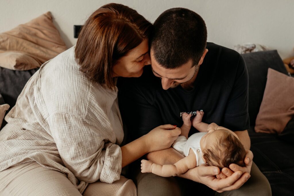 Mama und Papa sitzen auf der Couch. Der Papa hat das Baby auf dem Rücken liegend in seine, auf den Beinen abgestützten, Unterarmen liegen. Die Mama kuschelt ihr Gesicht an den Kopf des Mannes.