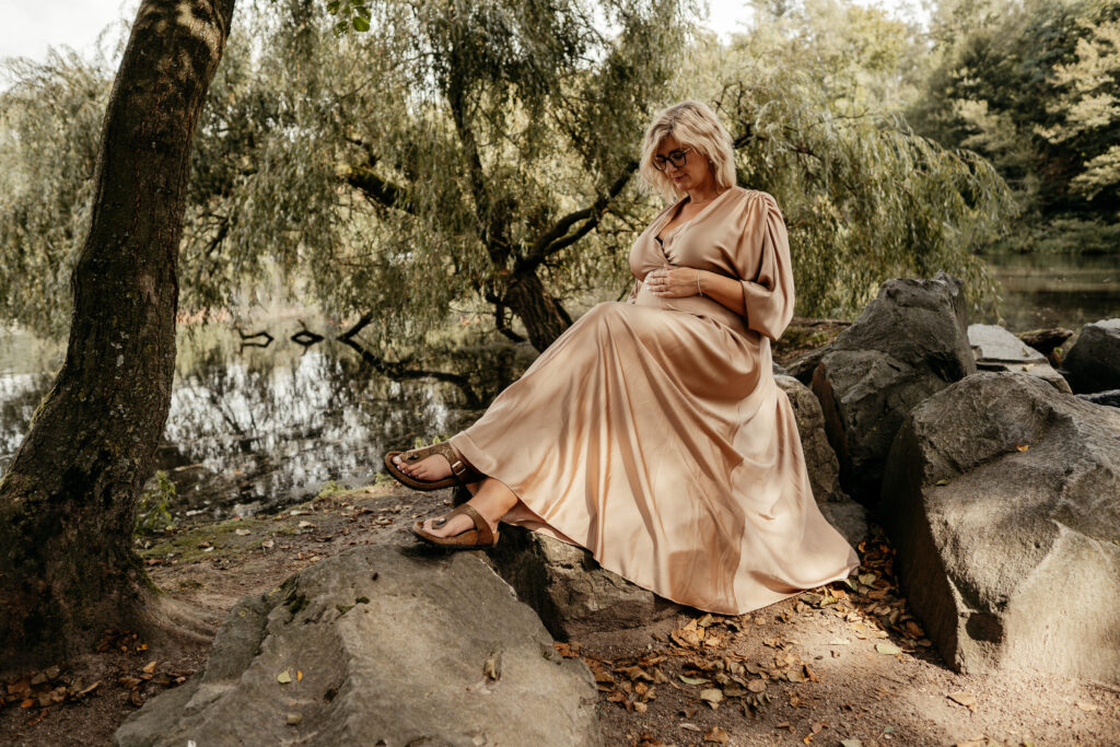 Schwangere Frau sitzt in einem beigen Satinkleid auf einem großen Stein unter einer Trauerweide und schaut auf ihren Bauch.