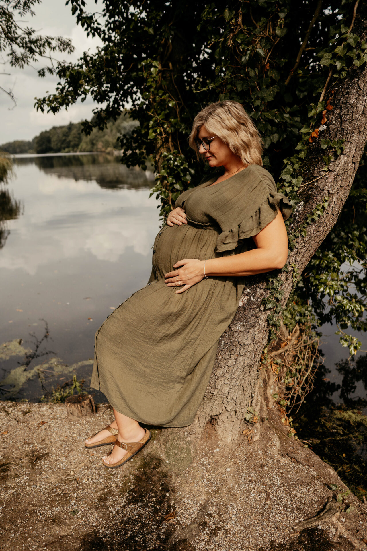 Schwangere Frau in einem Grünen langen Kleid, lehnt an einem Baum und hält liebevoll ihren runden Bauch.