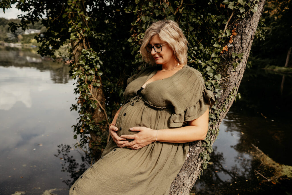 Schwangere Frau in einem Grünen langen Kleid, lehnt an einem Baum und hält liebevoll ihren runden Bauch.