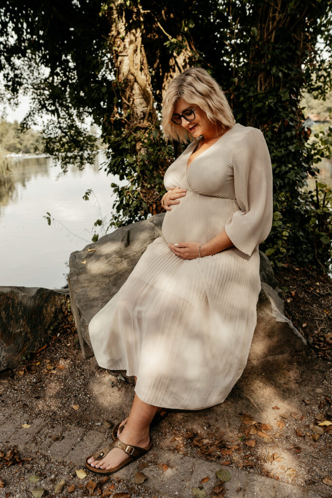 Schwangere Frau in einem beigen Plisseekleid, sitzt auf einem großen Stein und schaut verträumt auf ihren Bauch