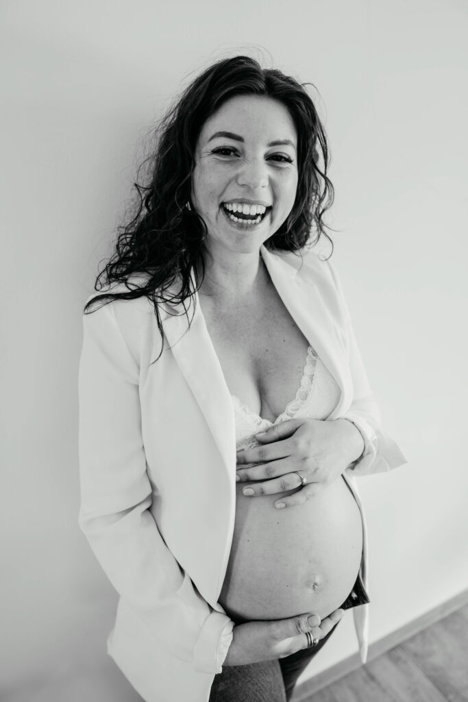 Schwangere Frau bei einem Babybauchshooting Zuhause. Die Schwangere lacht herzhaft in die Kamera während sie ihren nackten Bauch hält.