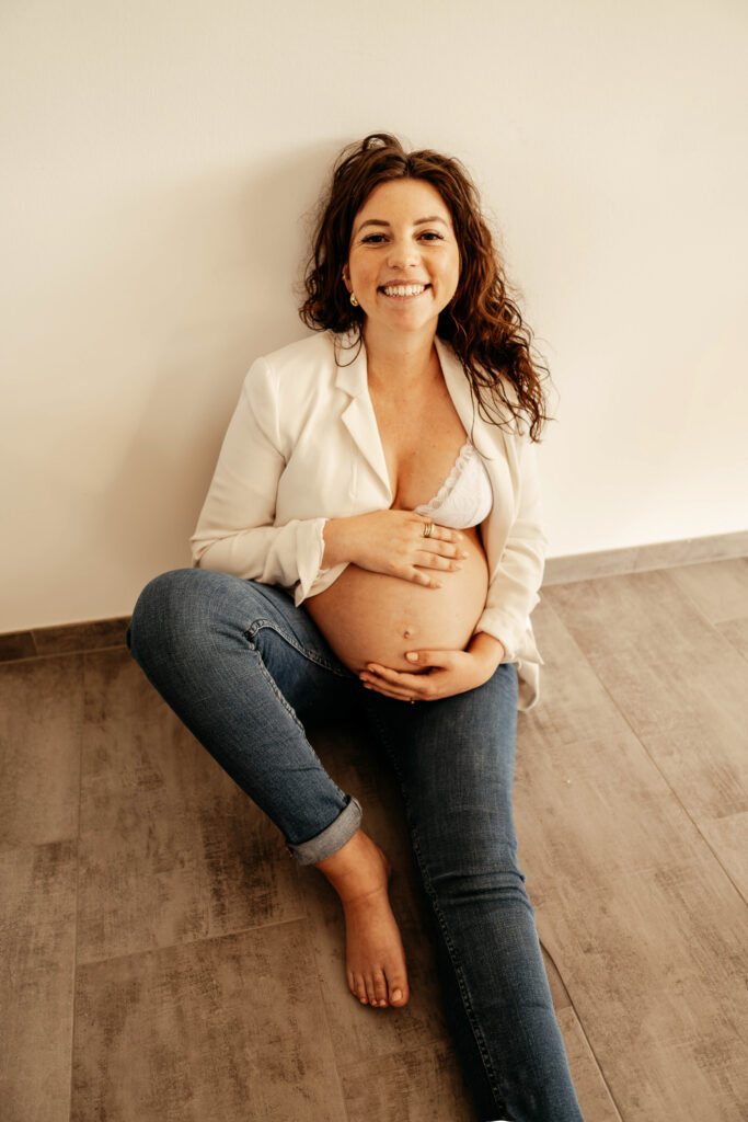 Schwangere Frau bei einem Babybauchshooting Zuhause. Sie sitzt auf dem Boden und lächelt in die Kamera
