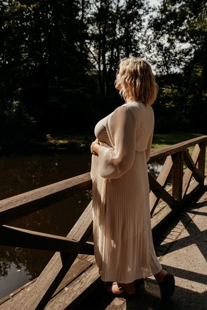 Schwangere Frau steht auf einem Steg am Geländer und schaut raus aufs Wasser. Dabei hält sie ihren Babybauch