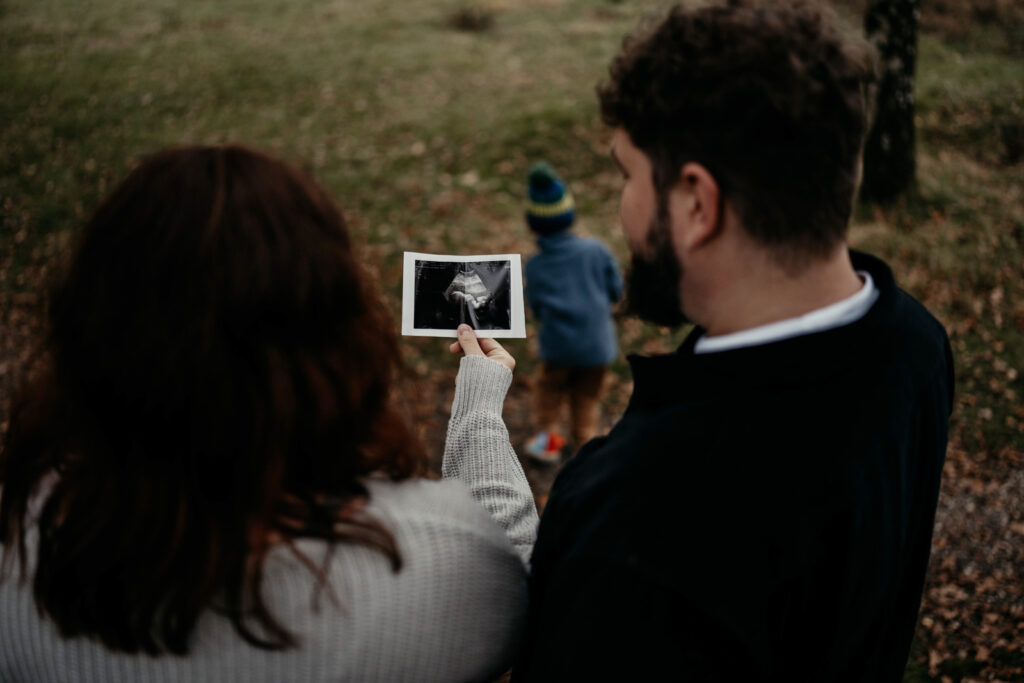 Ein Pärchen von hinten, zwischen den Köpfen der beiden kann man das Ultraschallbild sehen, welches die Mama in der Hand hält. Der dreijährige Sohn spielt im Hintergrund.