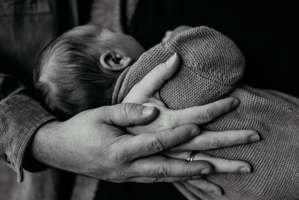 Eine schwarz-weiß Aufnahme, die den Rücken und den Hinterkopf eines neugeborenen zeigt das von der Mama mit der Hand am Rücken gehalten wird. Die Hand des Vaters liegt auf der Hand der Mutter.