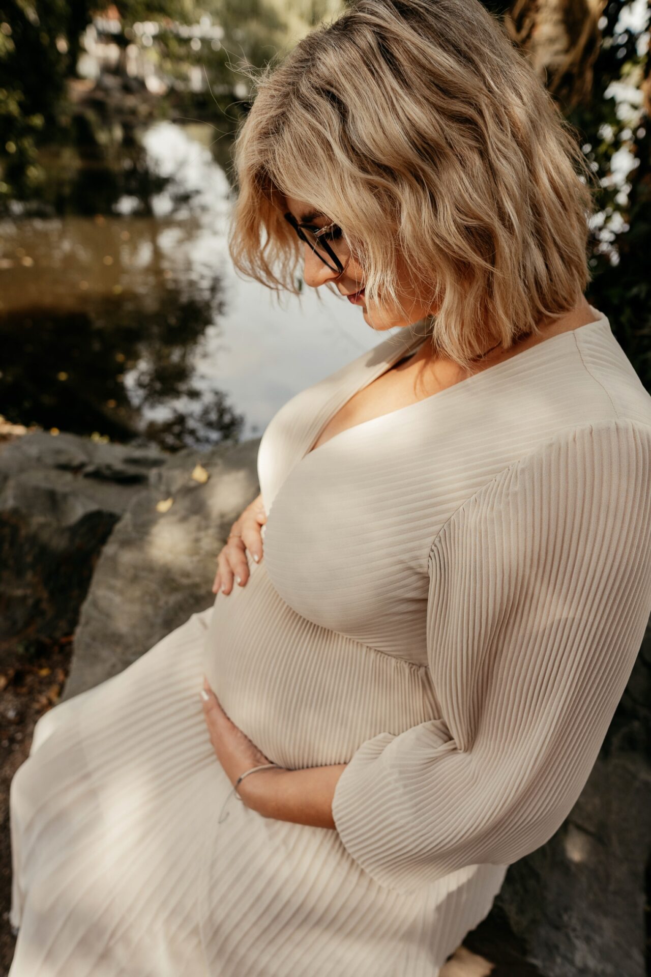 Frau mit Babybauch in beigem Kleid. Lehnt an einem Baum und hält liebevoll ihren Bauch. Babybauchfotografie in Brüggen