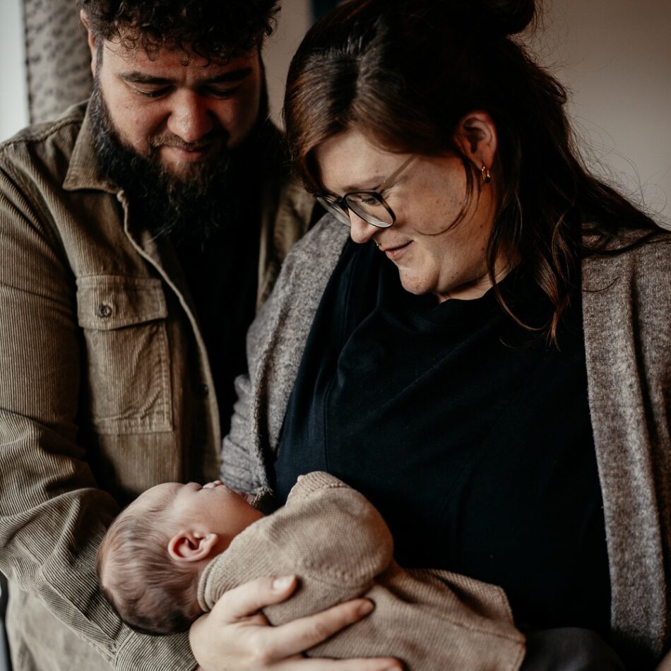 Foto von einem Neugeborenen-Homeshooting. Die Eltern halten gemeinsam das Baby im Arm und schauen es an.